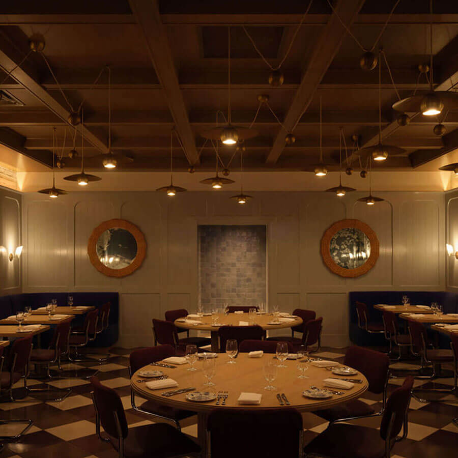 Warm main dining room with round wood tables set for service, burgundy chairs, navy banquettes, paneled gray walls with two round mirrors, checkerboard floor, and a coffered ceiling dotted with brass pendant lights